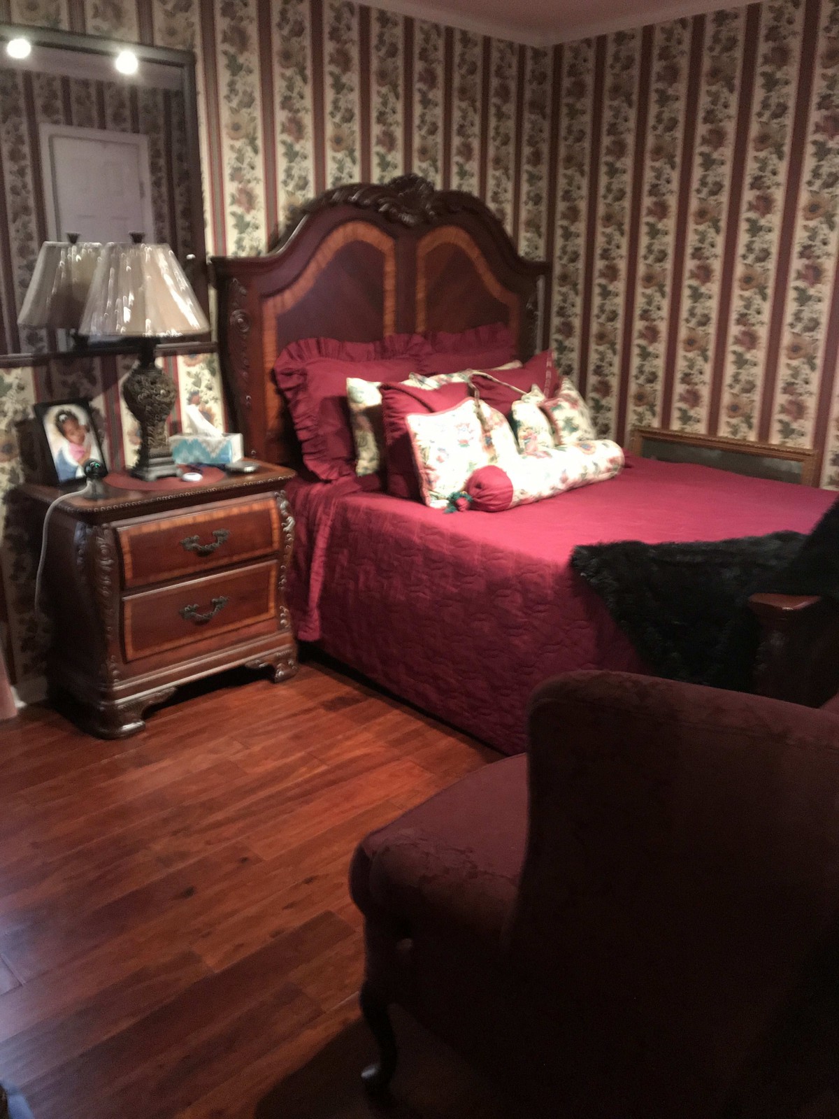 A cozy vintage-style bedroom with a wooden bed frame, matching nightstand, and floral wallpaper.
