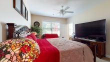 A cozy bedroom with a red-themed bedspread, large TV, and ceiling fan.