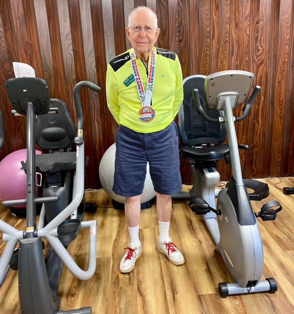 An elderly man standing proudly wearing a medal in a gym, with exercise bikes on either side.