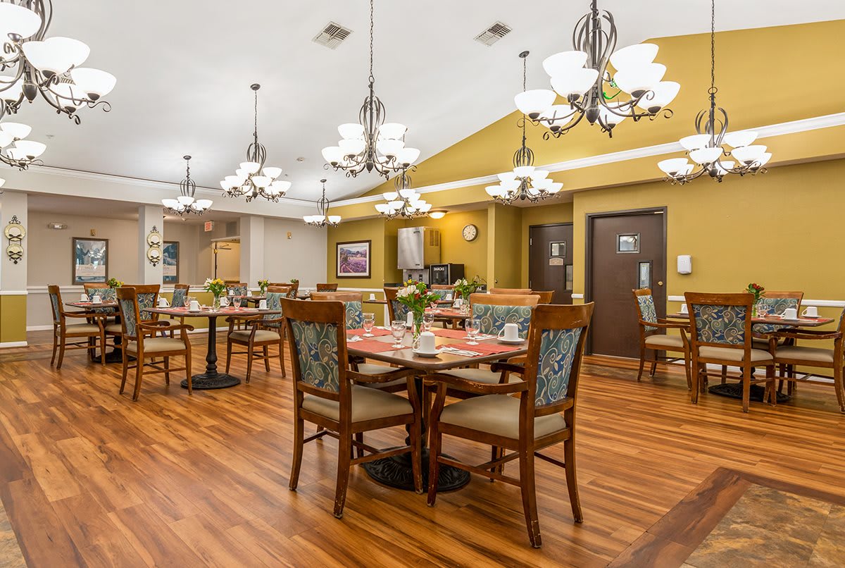 A warmly lit dining area with wooden floors, elegant chandeliers, and neatly arranged tables and chairs.