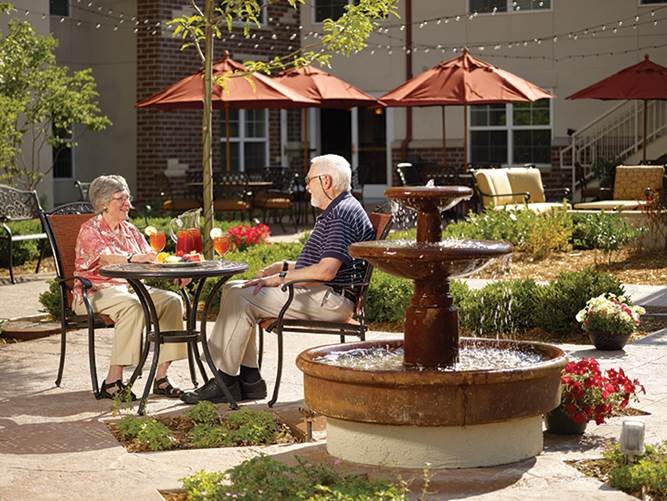 Two seniors sitting by a garden fountain under red umbrellas.