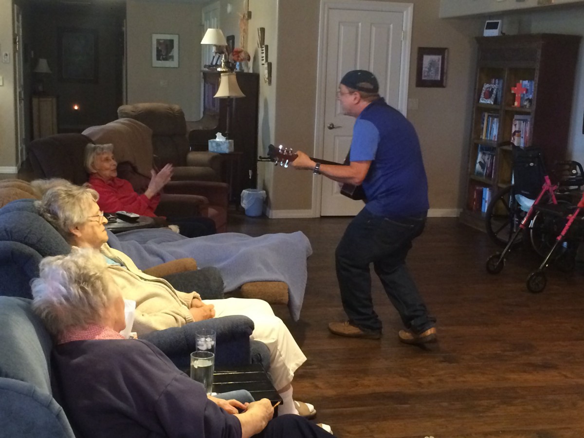 A person playing guitar for an audience in a cozy living room setting.