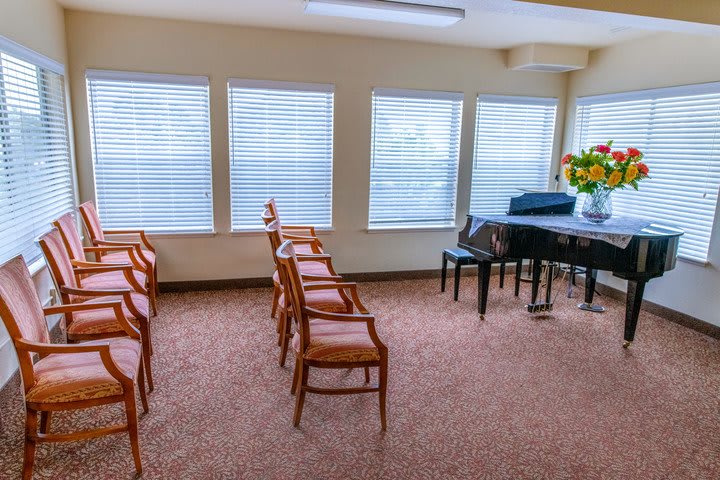 A small room with a piano, several chairs arranged in rows, and large windows allowing natural light to fill the space.