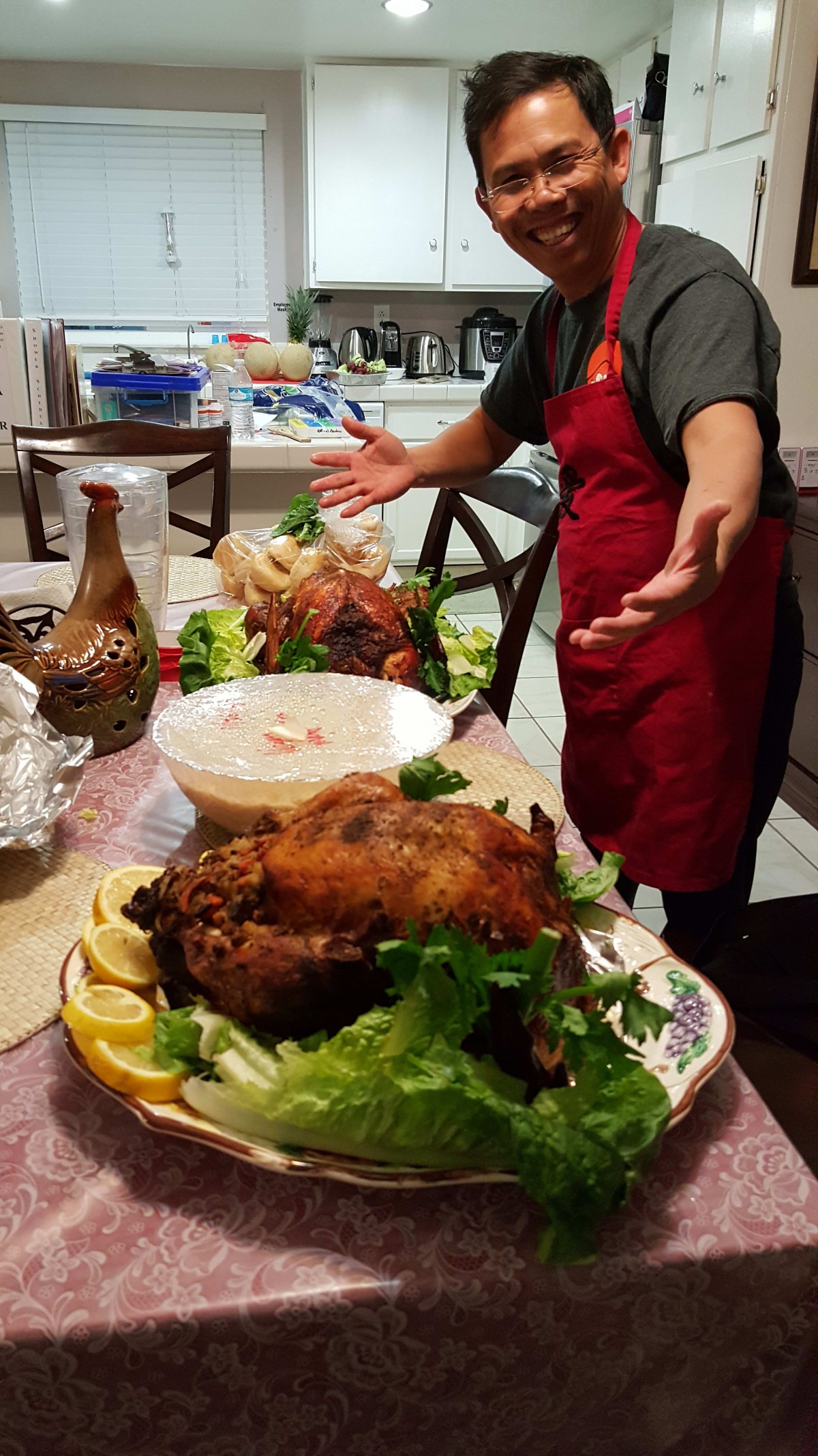 A person in a red apron presenting a table set with a roasted turkey and various dishes.