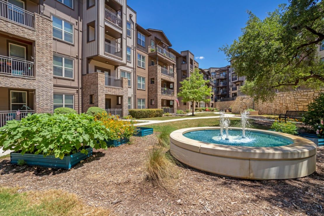 A modern apartment complex with a landscaped garden and fountain under a clear blue sky.