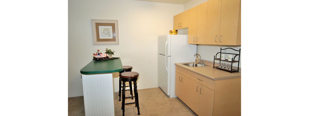 A compact apartment kitchen with wooden cabinets, a white fridge, and bar stools at the counter.