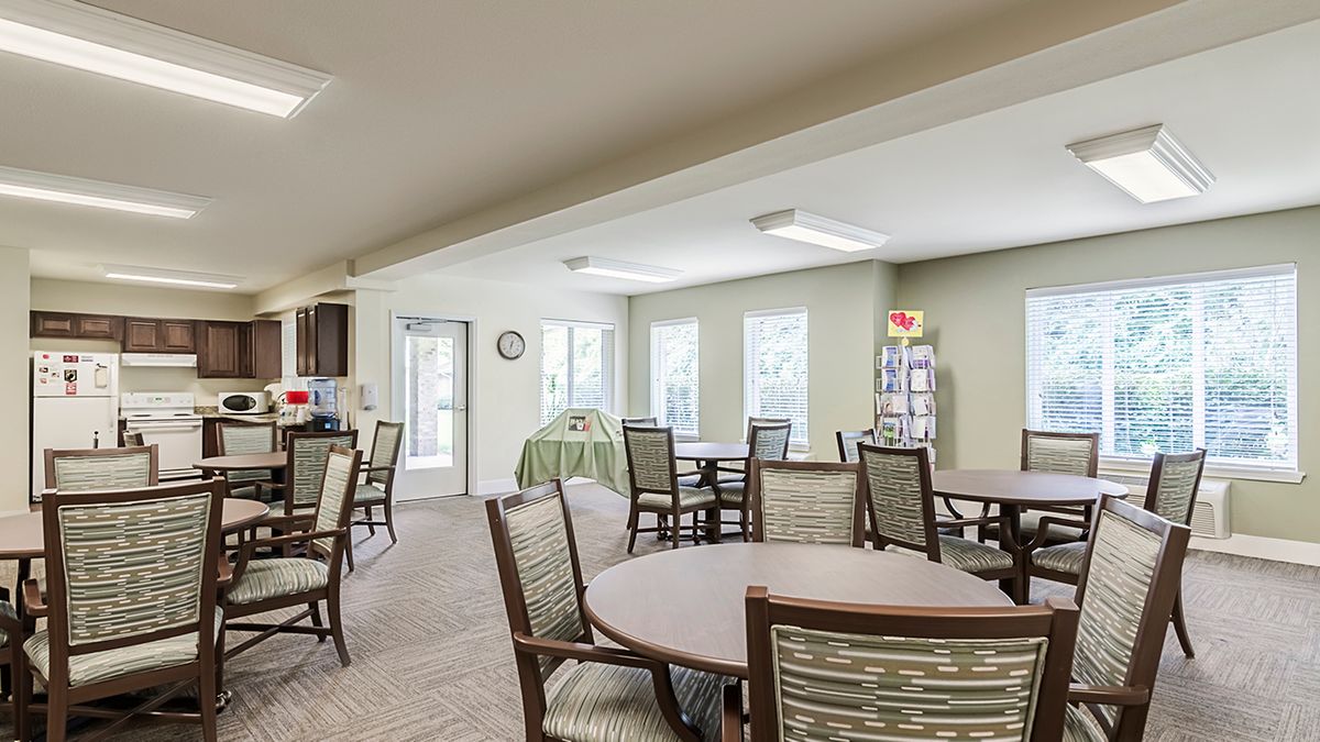 A spacious community dining area with several tables and chairs, well-lit by natural light through multiple windows.