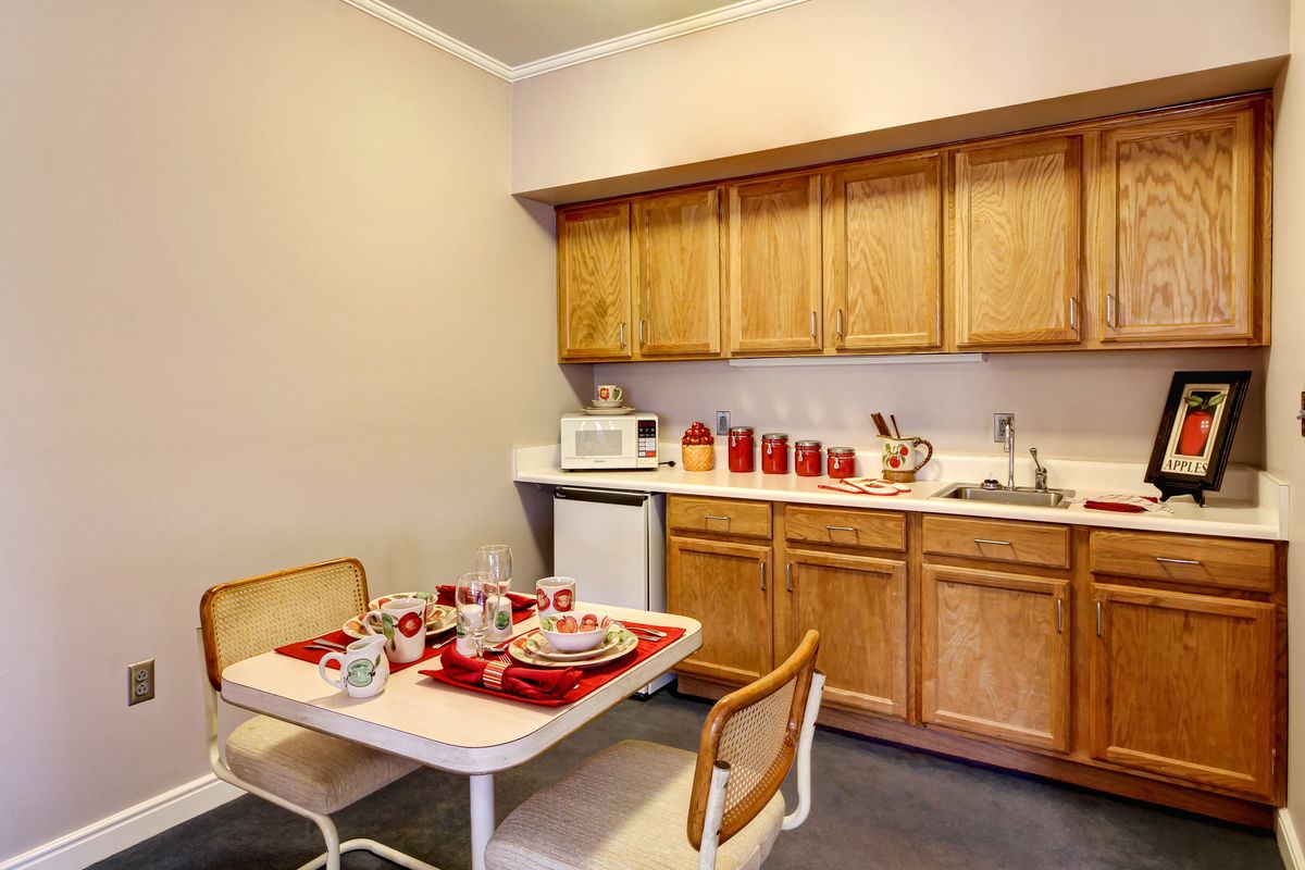 A cozy kitchen with a dining area, featuring wooden cabinets, a microwave, and a table set with red-accented tableware.