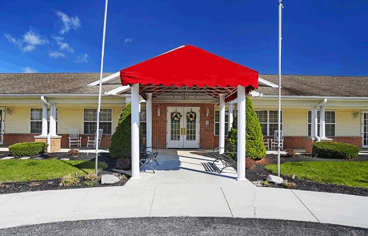 A building entrance with a red awning, supported by white pillars and surrounded by well-maintained landscaping.