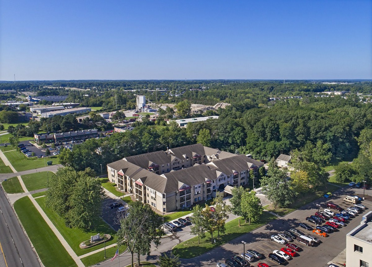 Aerial view of an apartment complex surrounded by trees and a parking lot in a suburban area.