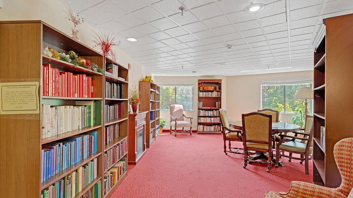 A cozy library room with bookshelves, a reading table, and chairs on a red carpeted floor.