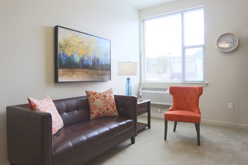 A cozy living room with a brown sofa, orange chair, and a colorful painting on the wall.
