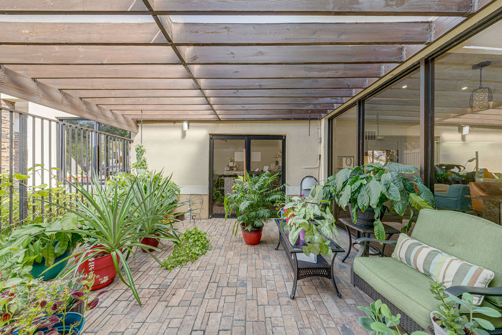A cozy patio with a variety of potted plants and comfortable seating under a glass roof.