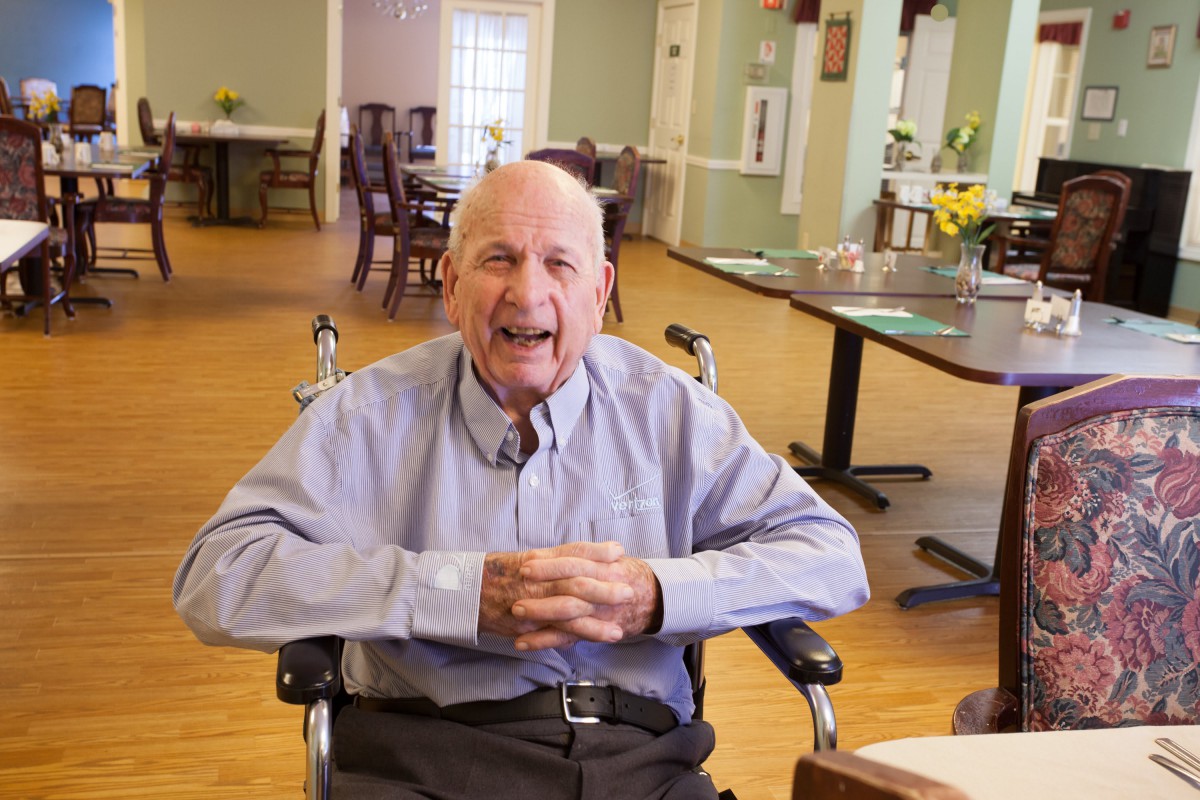 An elderly man sitting in a wheelchair, smiling indoors.