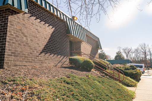 A side view of a brick building with sunlight casting shadows, featuring decorative overhangs and landscaped surroundings.