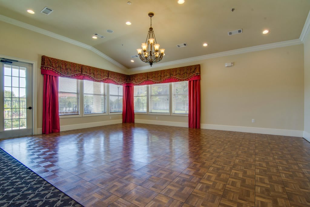 An empty room with hardwood flooring, large windows adorned with red curtains, and a chandelier hanging from the ceiling.
