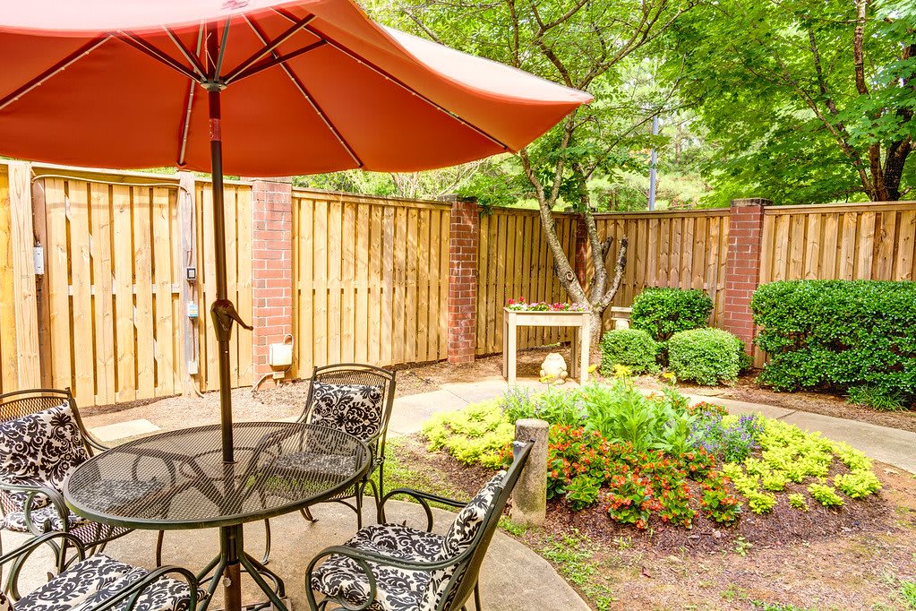 A cozy patio with a table and chairs under an orange umbrella, surrounded by a lush garden.