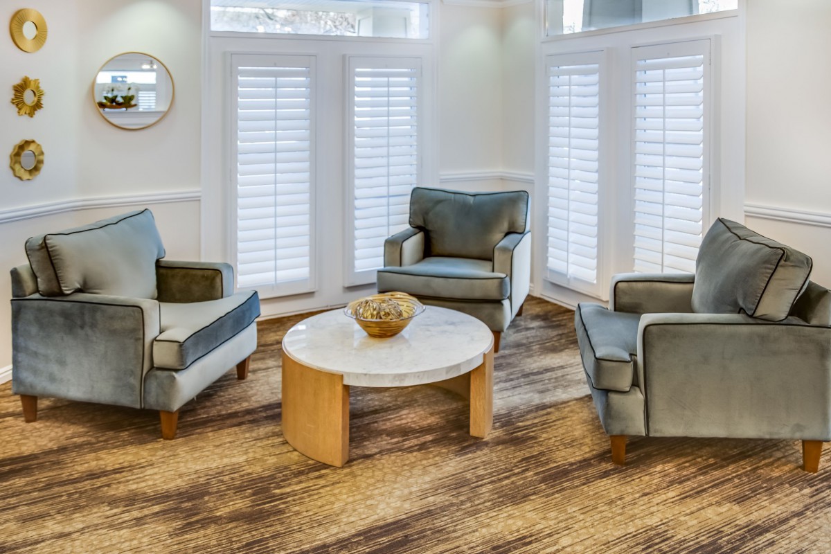 A modern living room with plush armchairs and a wooden coffee table, set against a backdrop of large windows with white shutters.