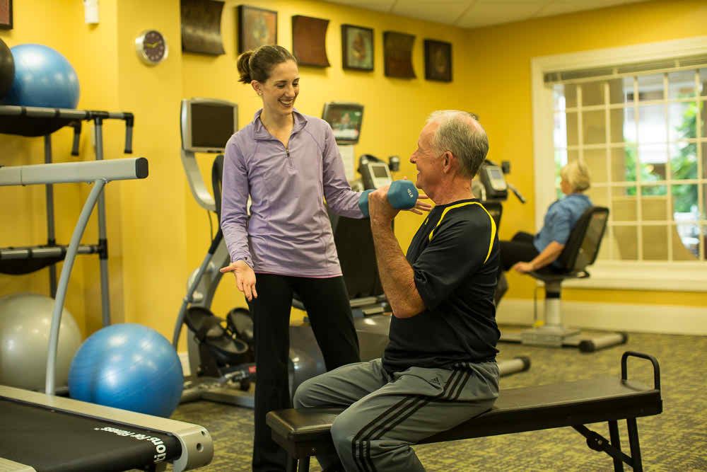 A personal trainer assists a senior man with weightlifting exercises in a gym setting.