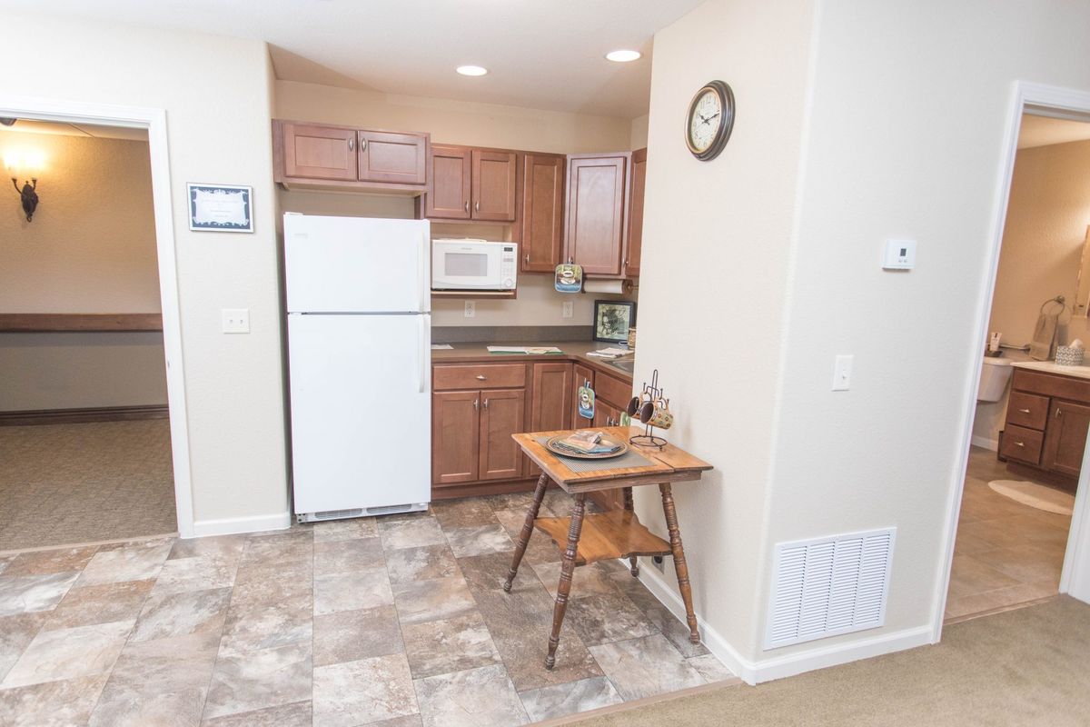 A small apartment kitchen with wooden cabinets, a refrigerator, and a microwave. A table and chair are set against the wall.