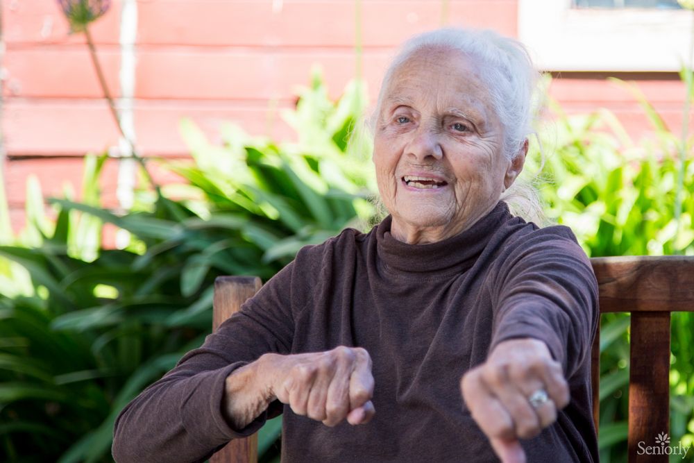 An elderly woman smiling while sitting outside surrounded by greenery.