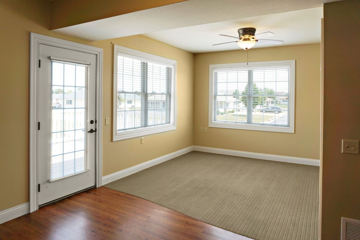 A bright empty room with a ceiling fan, large windows, and a mix of carpet and wood flooring.
