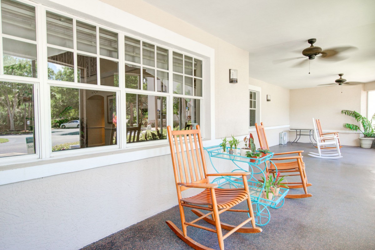 A cozy porch featuring two wooden rocking chairs and a glass table adorned with small plants, set against a backdrop of large windows.