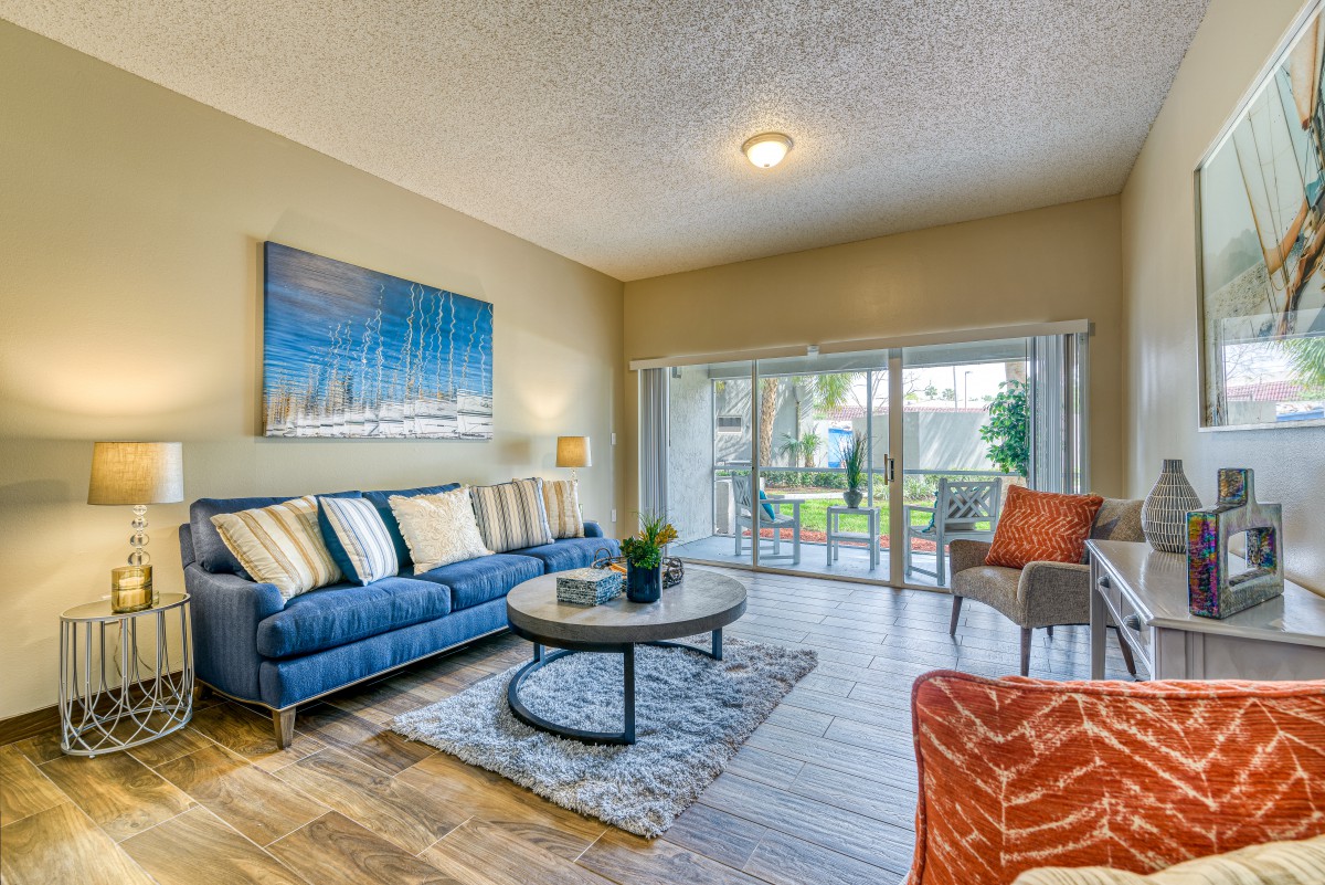 A modern living room with a comfortable sofa, coffee table, and vibrant artwork, illuminated by natural light from sliding glass doors leading to an outdoor area.