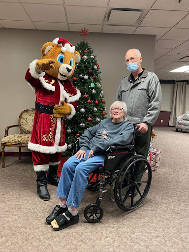 A festive moment captured with a cheerful mascot, an elderly individual in a wheelchair, and a companion, celebrating the holiday season.