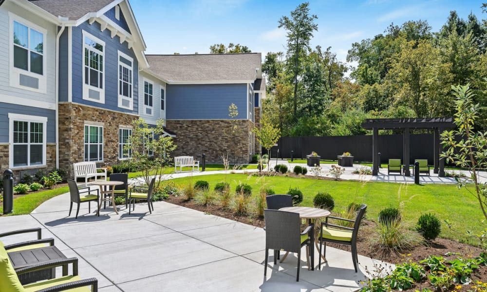 A sunny outdoor patio area with tables and chairs, surrounded by well-maintained landscaping and adjacent to a residential building.