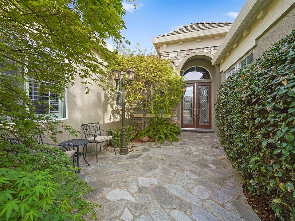 A charming house entrance with a stone path and outdoor seating area surrounded by lush greenery.