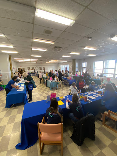 A busy community event taking place indoors, with people interacting at various tables.