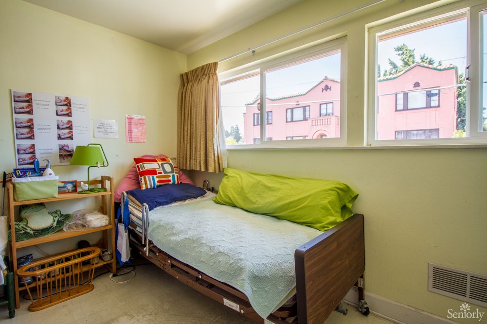 A cozy bedroom with a single bed by the window, featuring colorful bedding and a lamp on a small table.