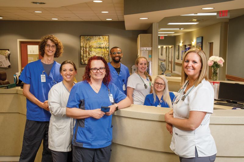 A group of healthcare professionals gathered at a hospital nursing station, smiling for a team photo.
