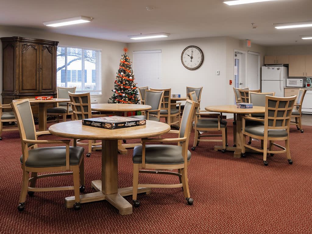 A community room with round tables and chairs, featuring a Christmas tree and a wall clock.