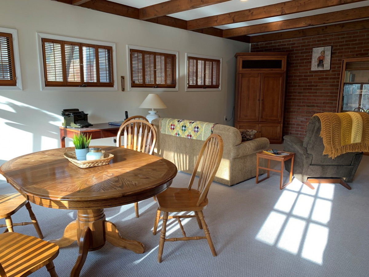 A cozy living room with a wooden dining table and chairs, a sofa, and ample natural light coming through the windows.
