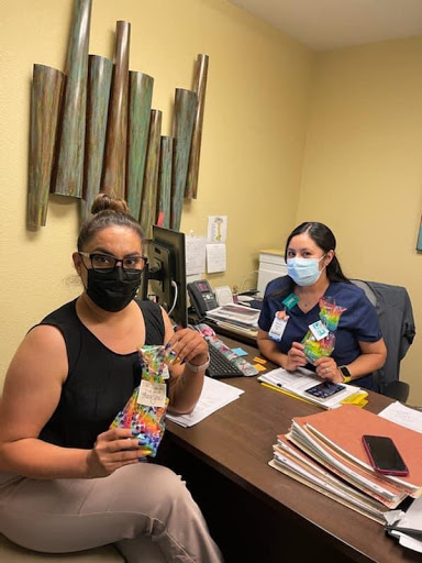 Two women in an office environment, both wearing masks and holding colorful items.