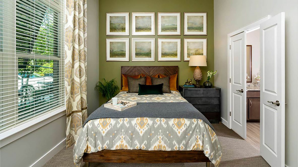 A cozy bedroom with a stylish interior, featuring a neatly made bed, framed artwork on the wall, and a side table with a lamp and plant.