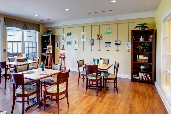 A cozy art gallery room featuring paintings on the wall, easels, and seating arrangements with tables and chairs.