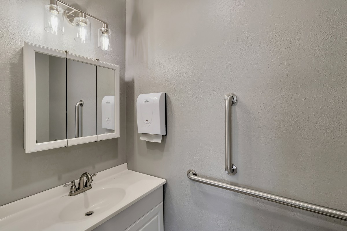 A modern bathroom with a sink, mirror, handrail, and soap dispenser.