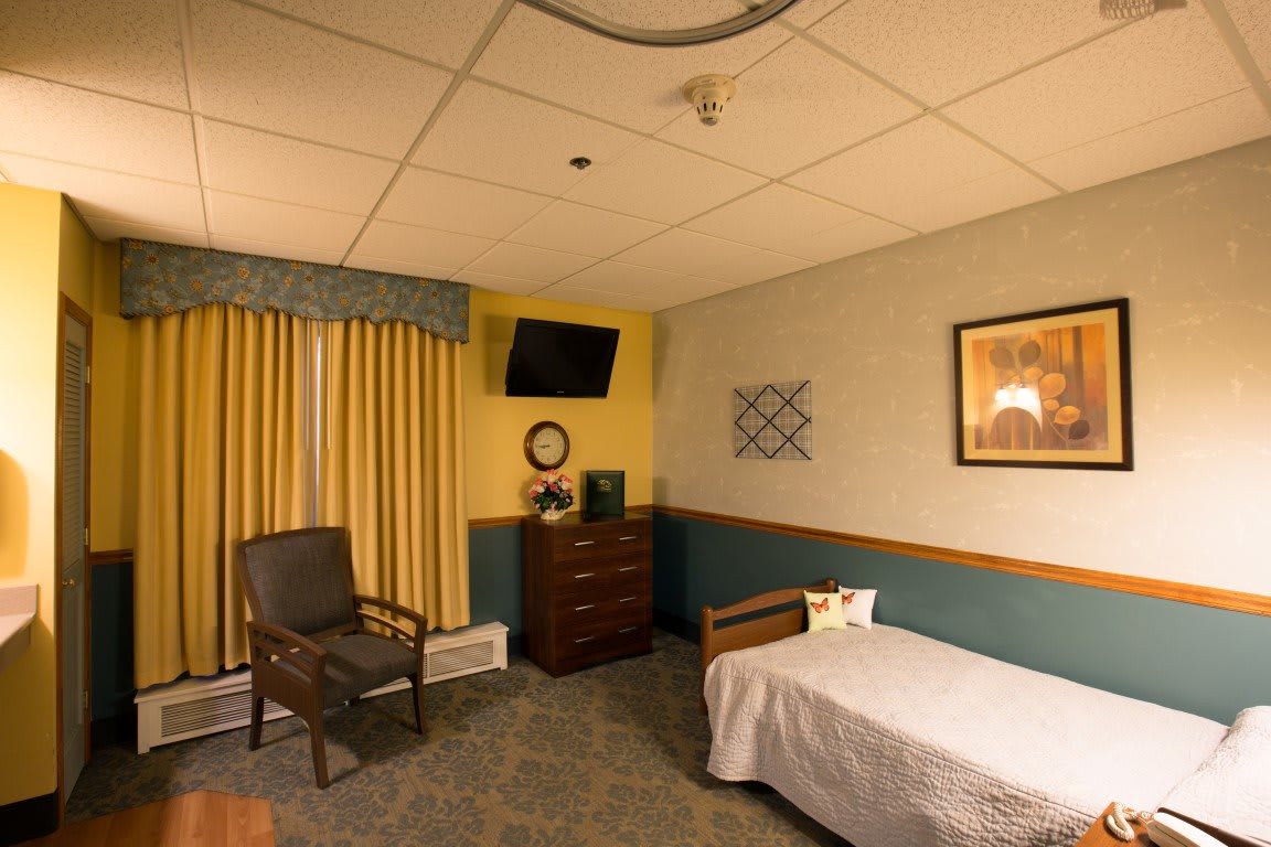 A cozy bedroom with a single bed, a chair, a dresser, and a wall-mounted television.