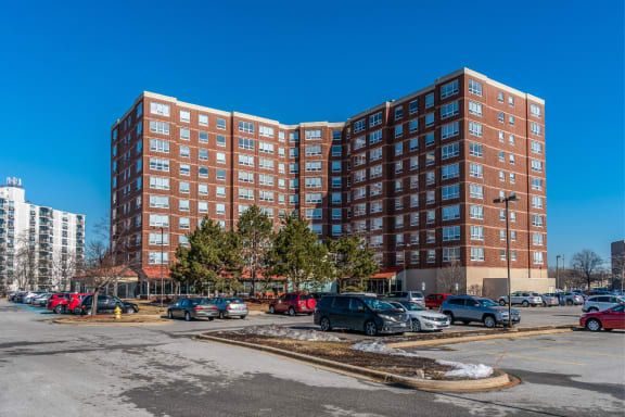 A large residential apartment building with a parking lot in the foreground under a clear blue sky.