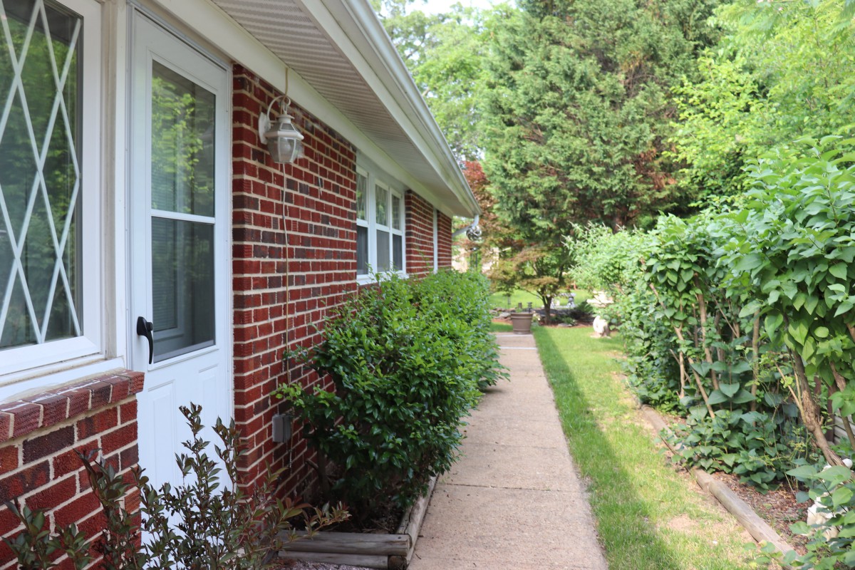 A brick house with a concrete pathway lined by bushes and greenery.