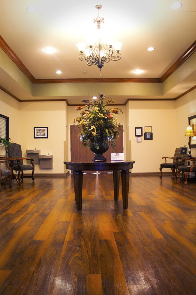 A warmly decorated lobby area with wooden flooring, a central table with floral arrangement, and seating along the walls.