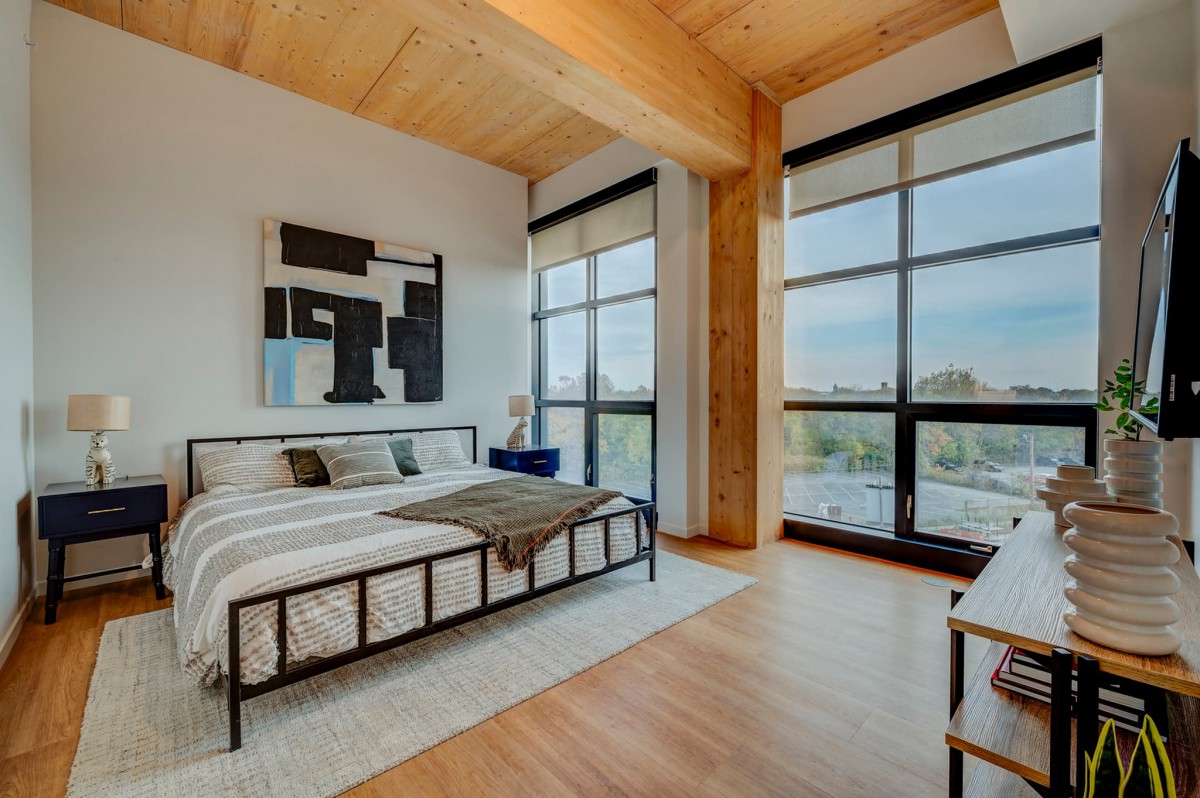 A modern, minimalist bedroom with large windows and wooden flooring, featuring a stylish bed and contemporary artwork.
