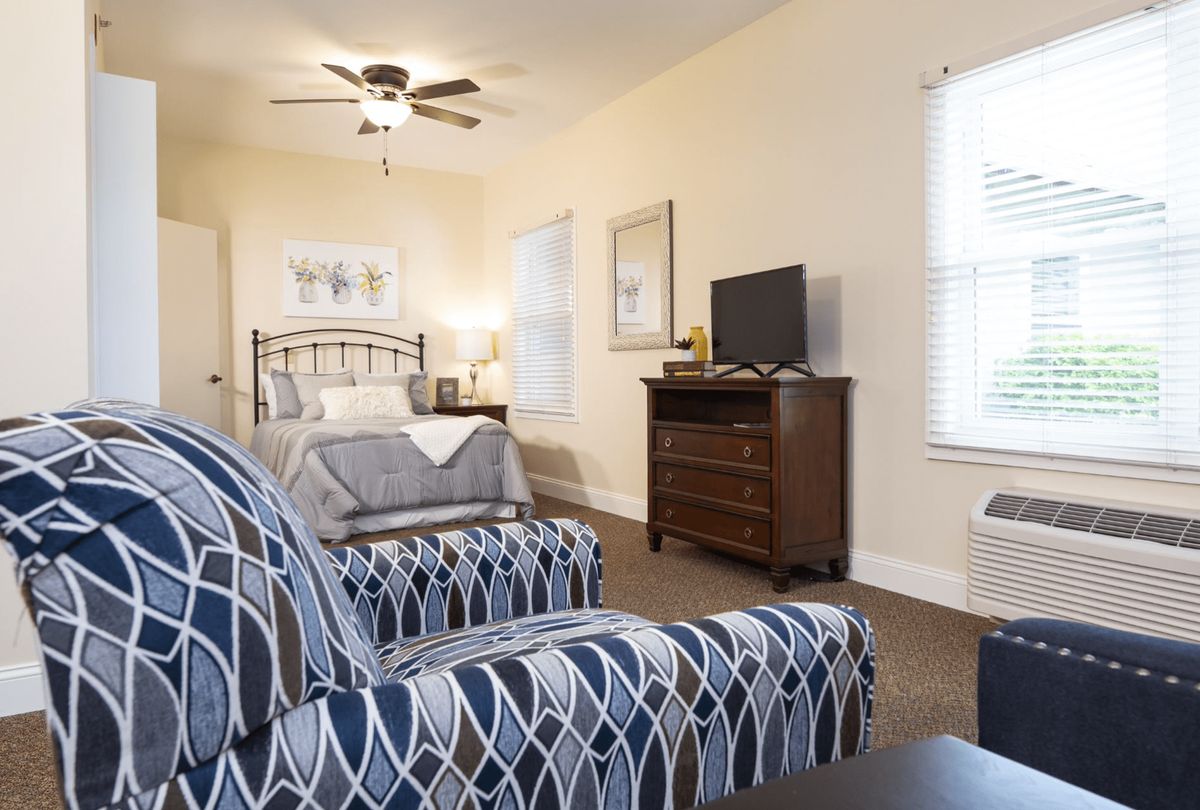 A cozy bedroom featuring a bed, patterned chair, dresser with a TV, and soft lighting.