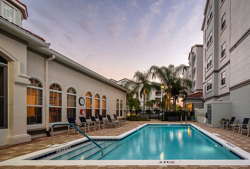 A tranquil outdoor swimming pool surrounded by palm trees and buildings, captured at sunset.