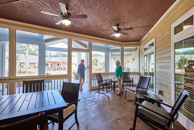 A screened porch with tables and chairs, featuring ceiling fans, overlooks a scenic outdoor area, with two people enjoying the view.