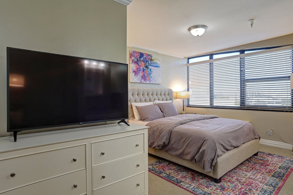 A cozy bedroom featuring a large bed, a TV on top of a dresser, and colorful artwork over the bed adjacent to a window.