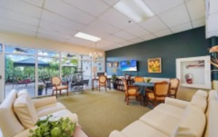 A cozy lounge area with comfortable seating, a television, artwork on the walls, and natural light streaming in through large windows.